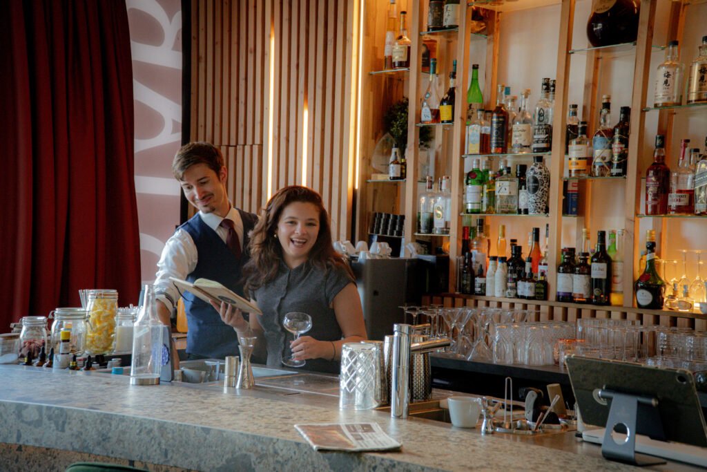 Un barman et une cliente sourient derrière le comptoir d'un bar bien achalandé.