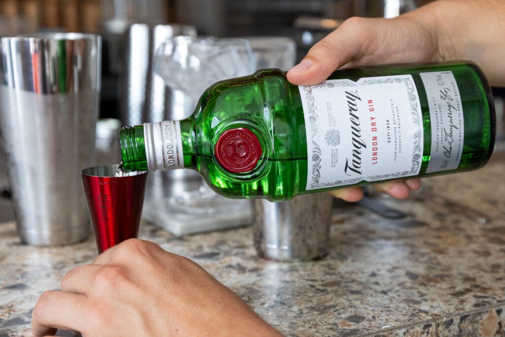 Wecandoo_Alexis-Perrichon_33_Cocktails_25082026-10 Versement de gin depuis une bouteille verte Tanqueray dans un doseur rouge sur un comptoir.