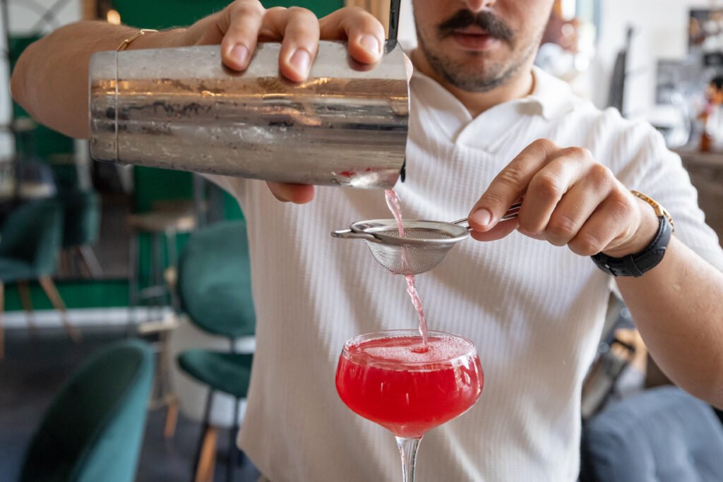 Wecandoo_Alexis-Perrichon_33_Cocktails_25082026-12 Un barman filtre un cocktail rose depuis un shaker dans un verre à pied.