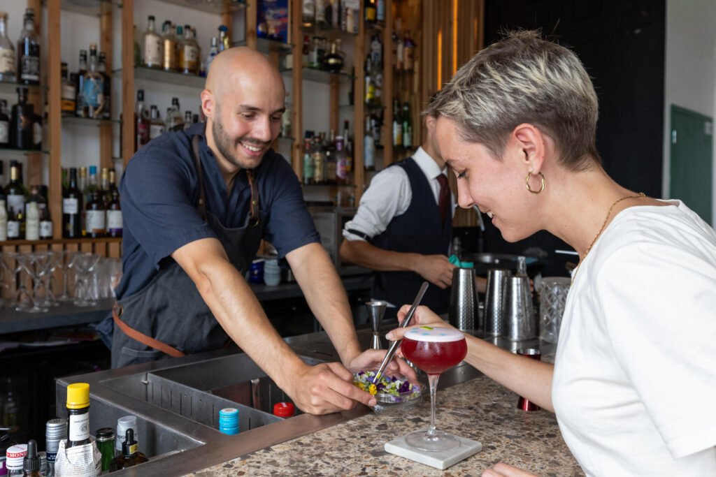 Un barman et une cliente posent une fleur sur un cocktail rouge au comptoir d'un bar.