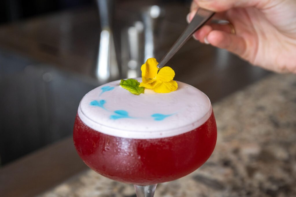 Gros plan sur la garniture d'un cocktail rouge avec une fleur jaune à l'aide d'une pince.
