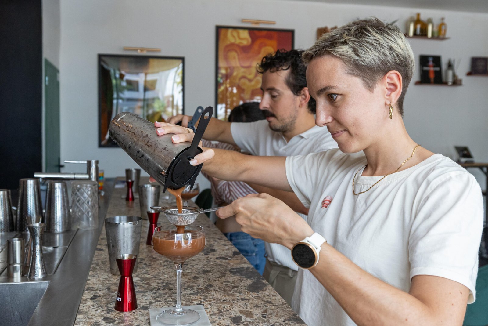 Une participante concentrée filtre un cocktail depuis un shaker dans un verre à pied, derrière le bar.