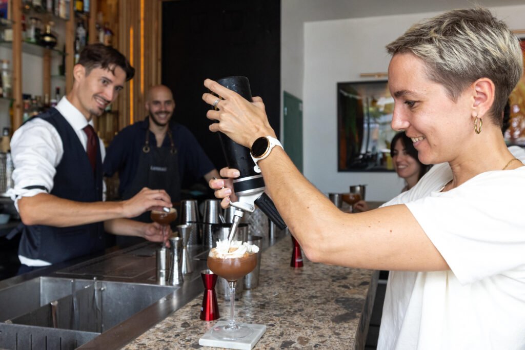 Une participante souriante applique de la crème sur son cocktail sur le comptoir d'un bar, encadrée par des barmans.